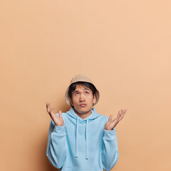 Vertical shot of hesitant Asian man spreads palms feels unaware and confused wears panama and blue...