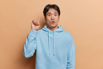 Horizontal shot of impressed Japanese man gestures upward with excitement indicating empty space for captivating ad content dressed in casual blue sweatshirt isolated over brown wall. Omg look there