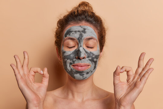 Peaceful woman keeps eyes closed and hands spread wide in zen gesture as she luxuriates in tranquil ambiance applies nourishing beauty mask isolated over beige background. Skin care concept.