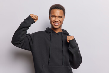 Handsome dark skinned adult man screams from happiness does winner gesture feels like winner dressed in casual black sweatshirt isolated over white background. People and celebration concept