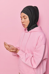 Sideways shot of calm young Muslim woman keeps hands clasped in traditional praying gesture has eyes closed wears casual hoodie and black veil isolated over pink background has faith in better