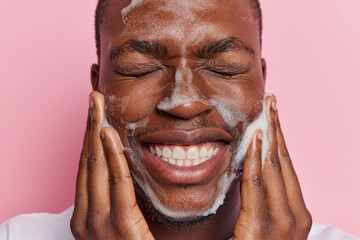 Close up shot of young African man exudes pure delight as he cleanses his face with soap pleased with condition of skin shows white teeth keeps eyes closed isolated over pink background. Daily hygiene - 614574075