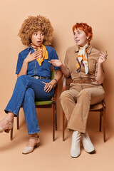 Full length shot of two women have pleasant talk surprised to find out news dressed in elegant costumes sit next to each other on chairs while waiting in queue isolated over brown background