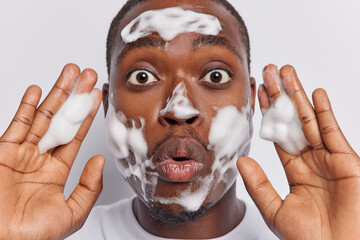 Close up shot of impressed dark skinned man takes care of condition of his skin keeps palms raised up washes face with soap bubble isolated over white background looks with refreshing contentment