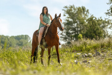 A young woman riding her bay brown horse in front of a rural summer landscape; female equestrian in summertime