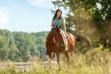 A young woman riding her bay brown horse in front of a rural summer landscape; female equestrian in...