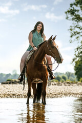 Equestrian and horse cooling down at a hot summer day: A young woman and her bay brown andalusian x arab horse gelding having fun in the water of a river outdoors