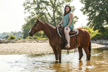 Equestrian and horse cooling down at a hot summer day: A young woman and her bay brown andalusian x arab horse gelding having fun in the water of a river outdoors