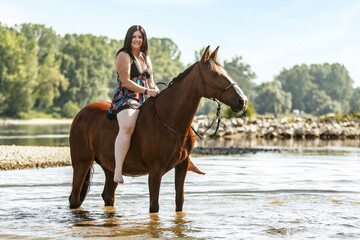 Equestrian and horse cooling down at a hot summer day: A young woman and her bay brown andalusian x arab horse gelding having fun in the water of a river outdoors