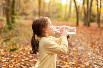 Little girl drinking water from a bottle in the autumn forest. Healthy lifestyle.