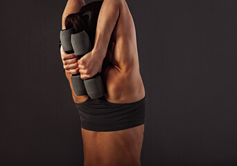 Fototapeta na wymiar Female muscular doing stretching and strength workout on the shoulders, blades and arms in sport bra holding dumbbells back behind herself standing on black background with empty space.