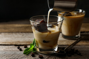 Pouring milk into glass of delicious iced coffee, mint and beans on wooden table