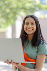 Beautiful Indian woman using laptop looking at camera on city street. Female preparing for exam