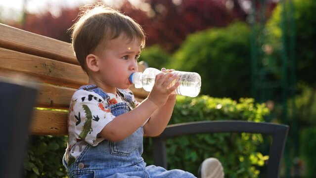 Cute Caucasian baby drinking water outside. Toddler sits on the bench holding a bottle.