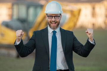 Successful construction business owner. Construction worker in suit and helmet near excavator....