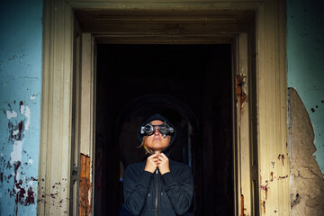 A blonde girl wearing steampunk glasses and a black hoodie in the doorway of an abandoned old building. A spectacular photo in the style of steam punk