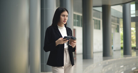 Obraz premium Young Asian business woman in suit holding mobile phone with takeaway coffee