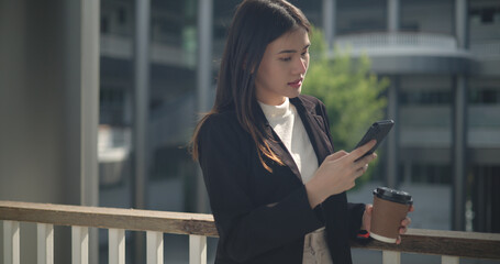 Fototapeta premium Young Asian business woman in suit holding mobile phone with takeaway coffee