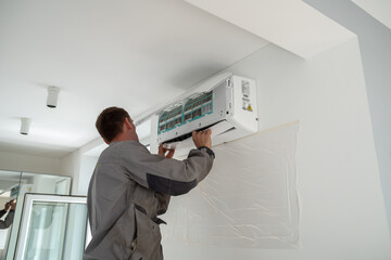 Male worker wearing uniform installing air conditioner in apartment during summer season, man technician standing indoors repairing HVAC system, checking and replacing AC filter