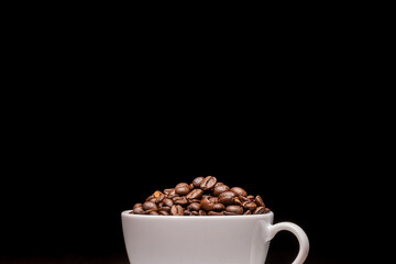 Coffee beans in a white coffee cup and many coffee beans placed around on a wooden table in a warm, light atmosphere, on a dark background, with copy space.