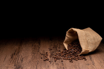 The many coffee beans and bag and scoop are placed around on a wooden table in a warm, light atmosphere, on dark background, with copy space.