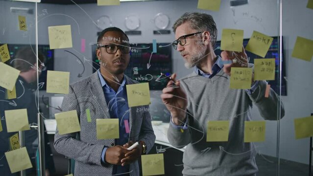 Multiracial financial analysts draw on glass wall, develop and analyze company crypto business strategy in investment bank office. Computers and big digital screens with real-time stocks on background