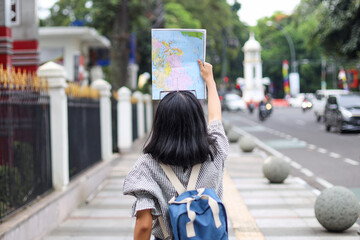 Back view of young woman with backpack goes down the street while holding the map. Travelling and...