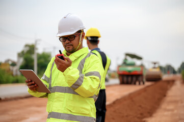 Asian surveyor engineers use tablet to check detail in job,  Control schedule of work of highway road construction projects with construction team and construct machinery in background.