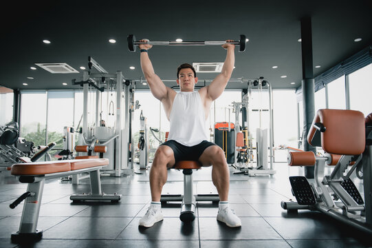Muscular bodybuilder Asian man sitting weight lifting bar in the gymnasium, exercise biceps with weight lifting bar workout. Fitness execute exercising building muscle concept.