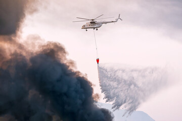 a firefighter emergency helicopter extinguishes a fire and sprays water from a basket over a column of black smoke over a city or wildfire