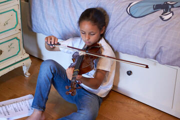 Violin lesson at home. Preteen girl is sitting on the floor of her bedroom while practicing violin. Horizontal composition, 