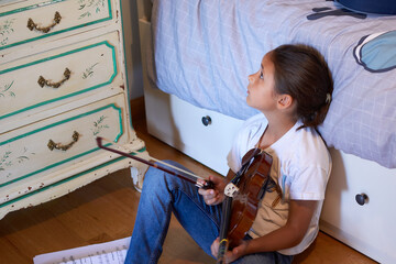 Violin lesson at home. Young girl sitting on the floor of her bedroom looking distractedly at the...