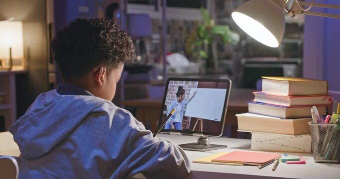 African american boy watching lesson online and studying from home at night. Smart primary school kid learning and taking notes while looking and making homework with teacher on laptop screen.