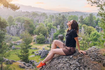 Obraz premium Happy traveler girl sitting on the rock cliff with view of stone remnants in Altinkaya, Turkey. Tourist attractions and destinations