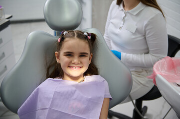 Close-up portrait of a happy little child girl smiles looking at camera, sitting in dentist's chair, receiving dental treatment in pediatric dentistry clinic. Dental health and oral hygiene concept