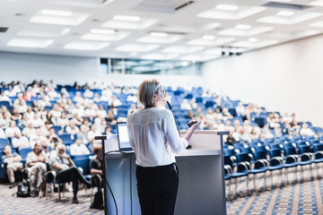 Female speaker giving a talk on corporate business conference. Unrecognizable people in audience at...