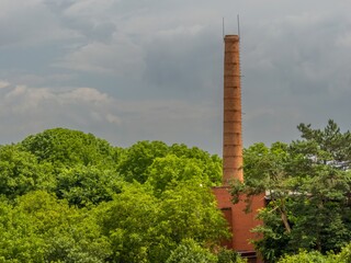Obraz premium high chimney of an old boiler house made of red brick surrounded by green trees against the background of clouds on a summer day