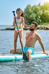 Father and daughter surfing on sup board and having funny time, family summer vacations.
