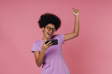 Excited woman making winner gesture while playing online game on mobile phone isolated over pink wall