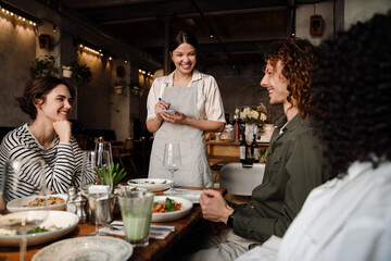Young waitress serving group of cheerful friends in restaurant