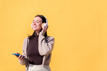 woman enjoying happy listening to music from mobile phone in headphones with a yellow wall in the...