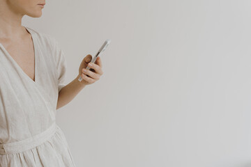 Mobile phone in female hands. Young pretty woman in neutral creamy beige linen dress holds cell phone over white wall. Social media, web surfing concept