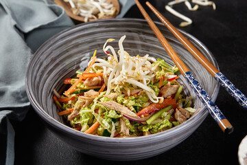 An appealing chicken salad with vegetables, Peking cabbage, and soy sprouts in an Asian style bowl, served with chopsticks on a black background. Accented with pink sea salt