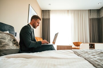 Sitting on the bed and working by using laptop. Businessman is indoors in the hotel room