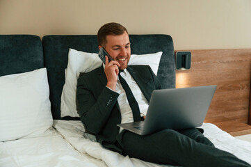 Smiling, lying down on bed and talking by phone. Businessman is indoors in the hotel room