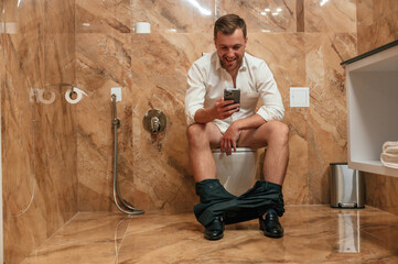 Watching funny content. A man in a white shirt is sitting on the toilet, holding a smartphone in...