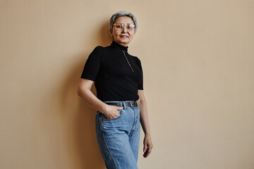 Waist up portrait of modern Asian senior woman looking at camera confidently standing against wall