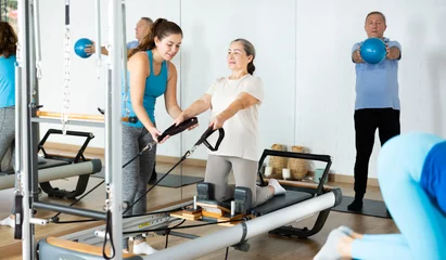 Fotobehang Persoonlijk Positive personal female trainer assisting elderly woman exercising on Pilates bed machine in rehabilitation center  © JackF