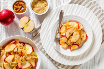fried onion and red apple slices on white plate and in bowl on white wood table with ingredients and spices, horizontal view from above, flat lay