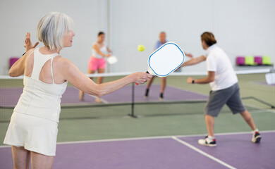 Rear view of fit elderly woman playing pickleball on indoor court, swinging paddle to return ball over net ..